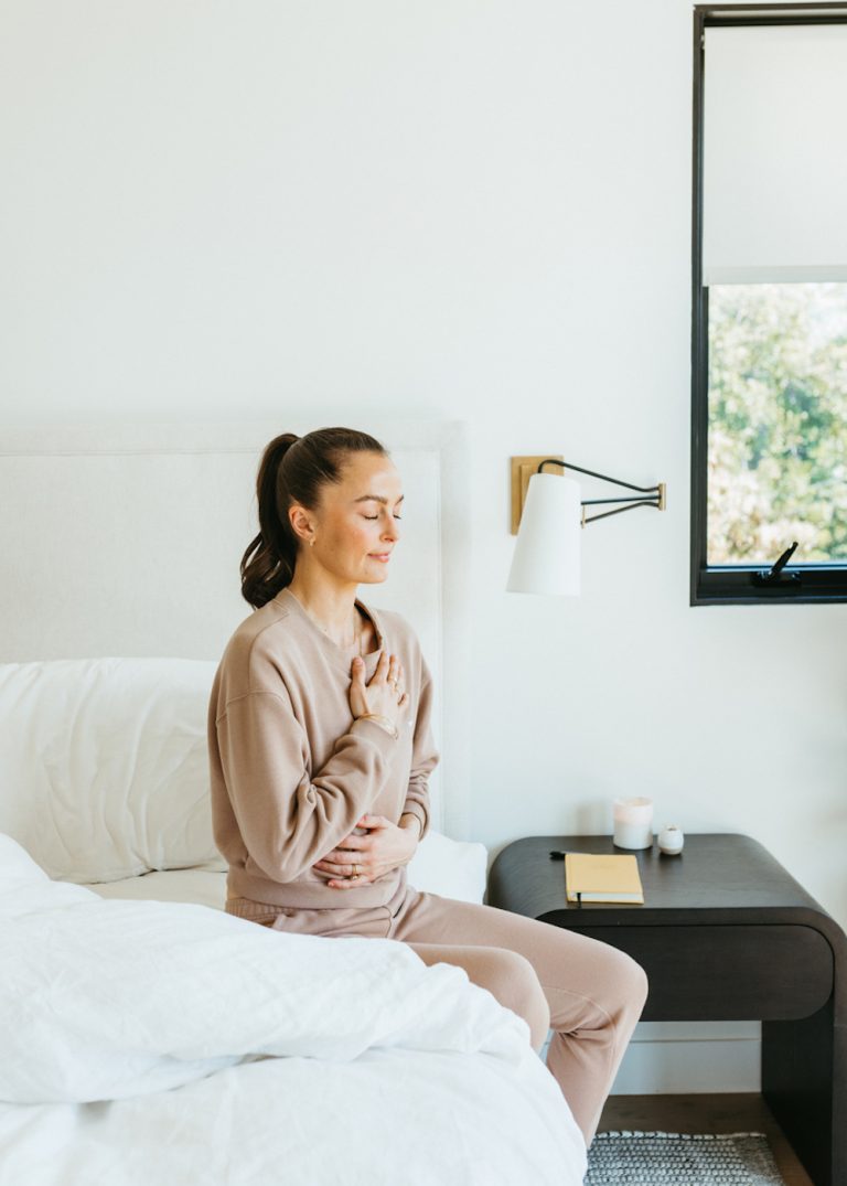 Brunette woman wearing brown sweatsuit meditating on bed.