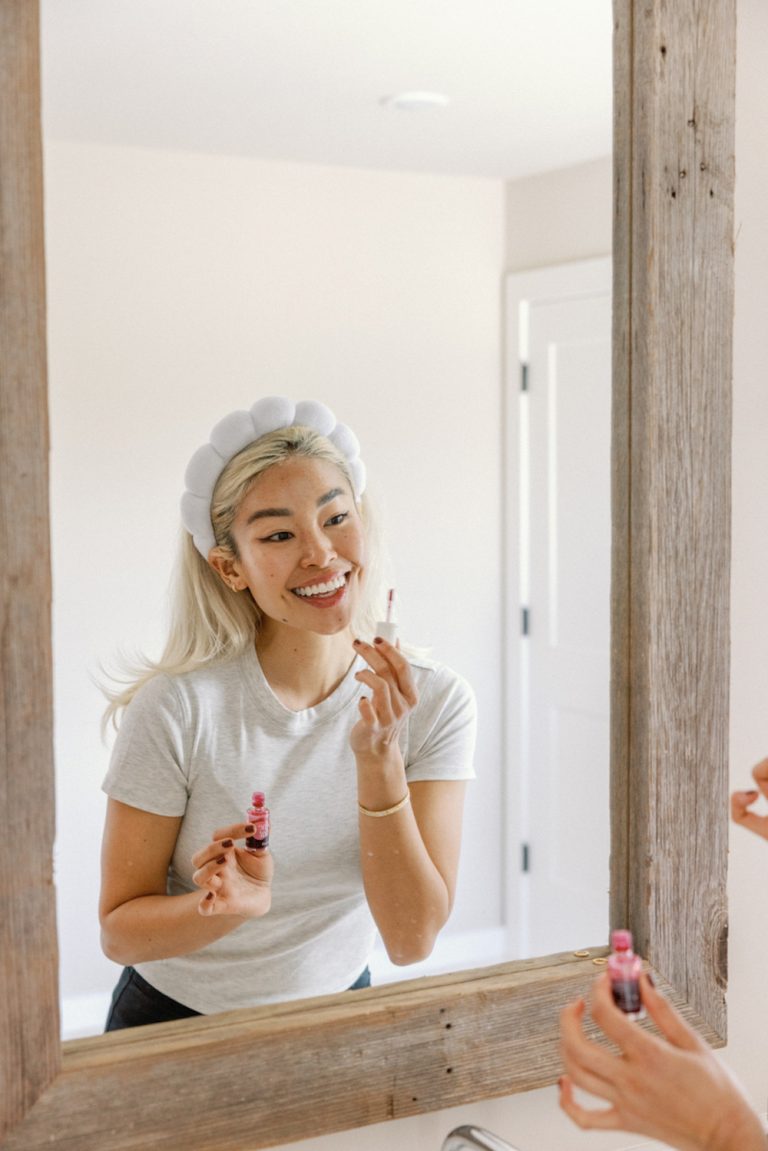 Blonde woman with headband applying lip gloss in the mirror.
