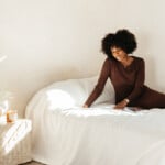 Black woman wearing brown sweater dress sitting on bed with white sheets.