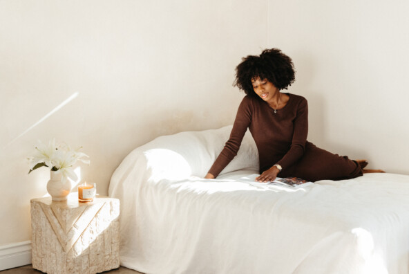 Black woman wearing brown sweater dress sitting on bed with white sheets.