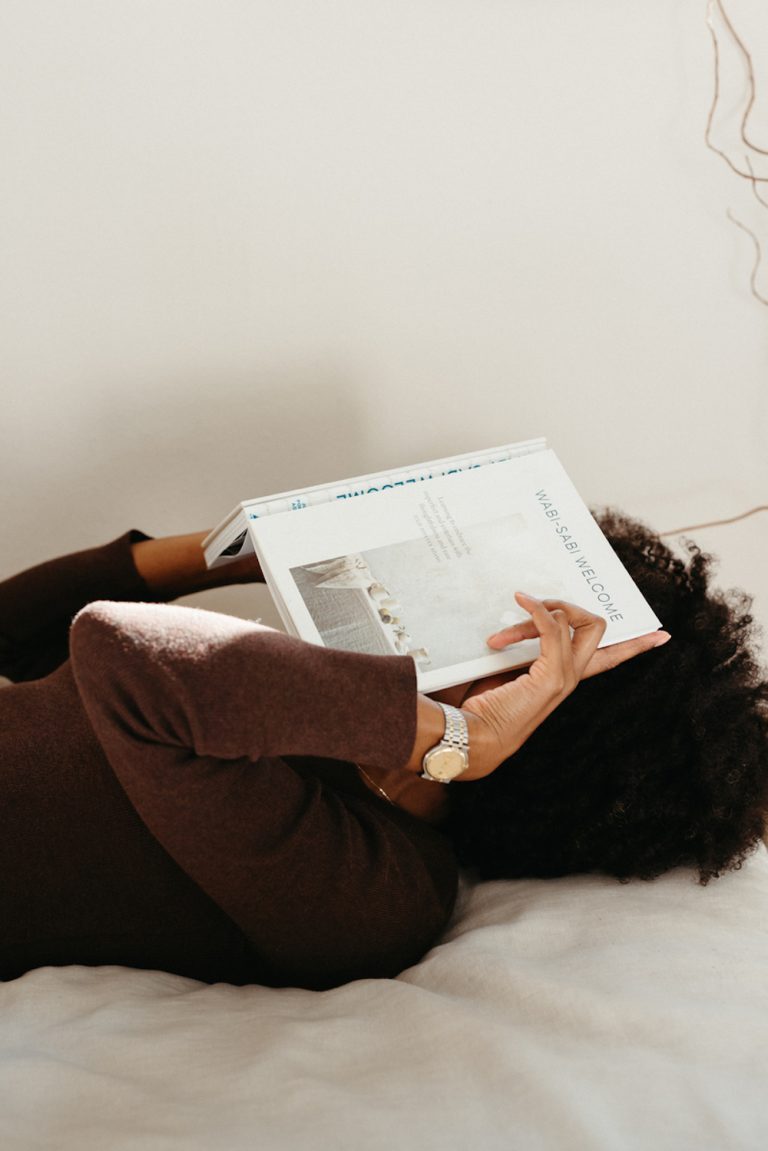 A woman in a brown sweater dress lies on a white sheet and covers her face with a book.