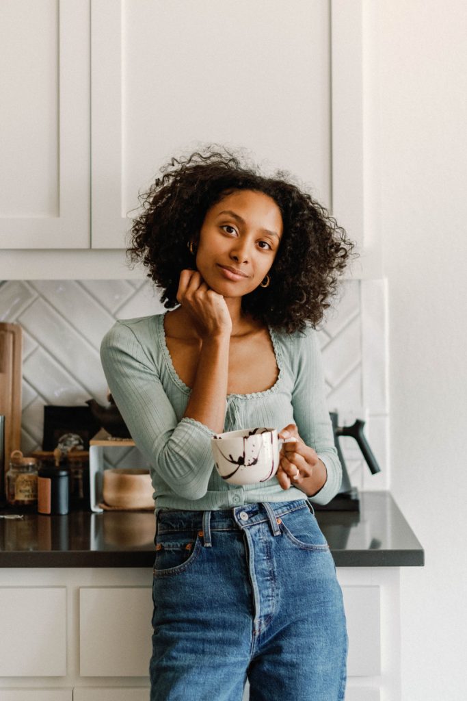 Black woman with dark hair leaning on kitchen countertop holding ceramic coffee mug.