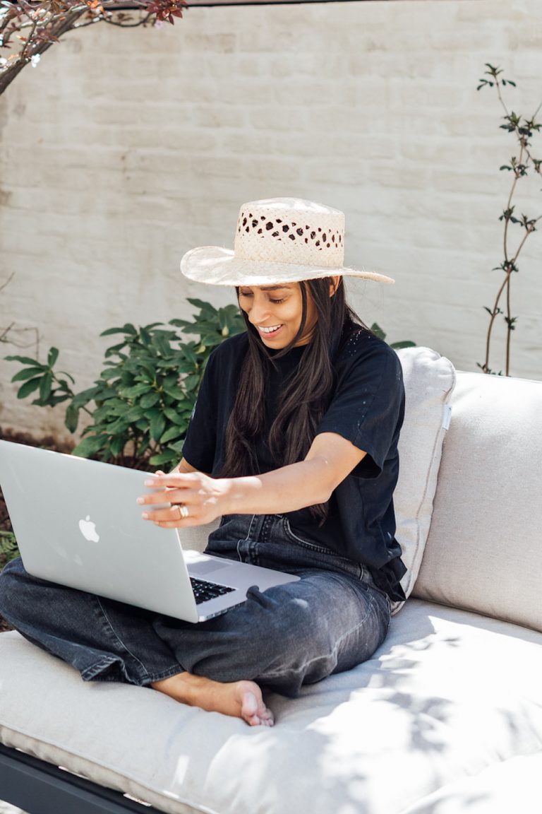 Brunette woman wearing sun hat working on laptop on outdoor patio.