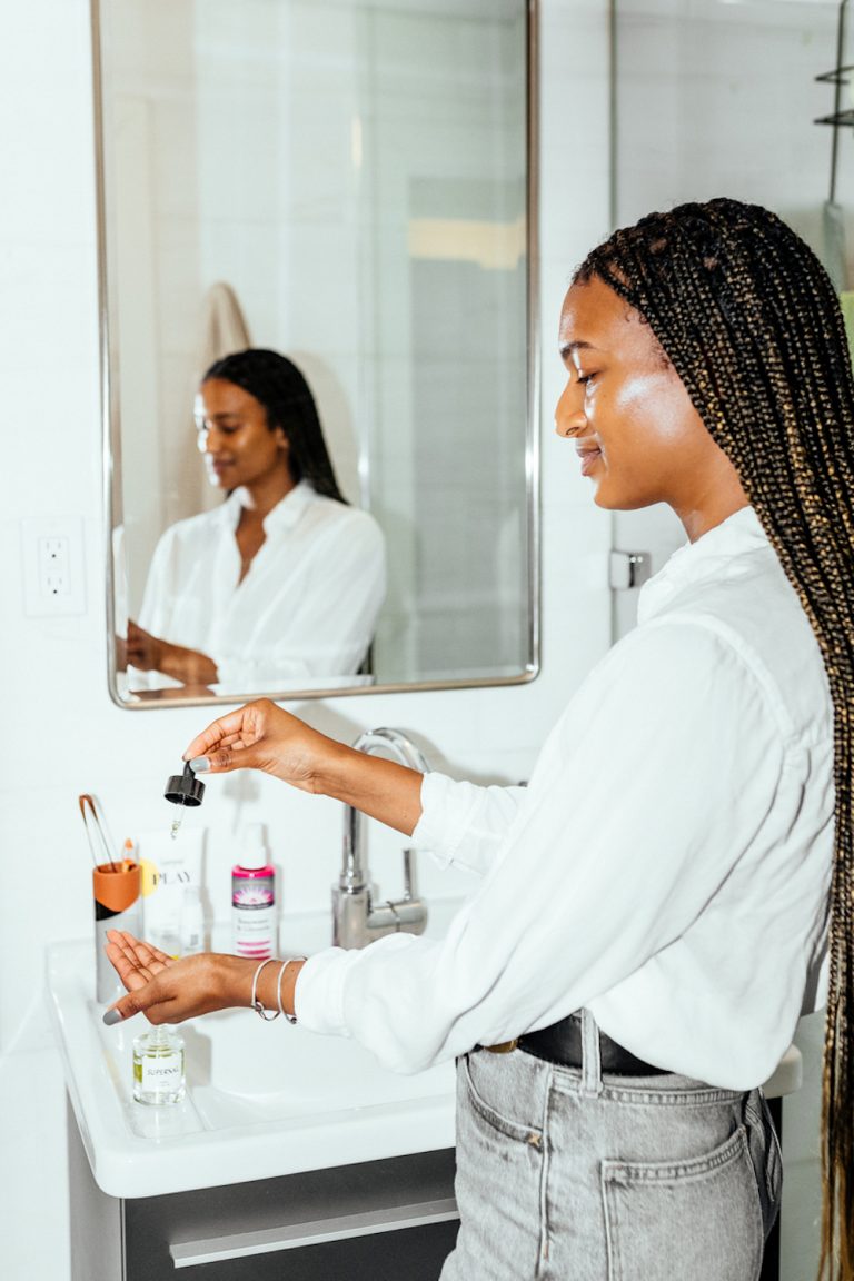 Black woman with long braids applying skincare in bathroom.