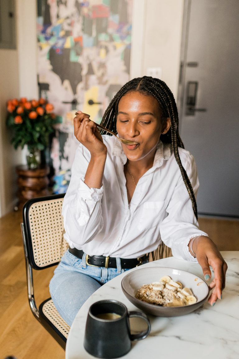 Woman eating bowl of oatmeal with bananas at dining room table.