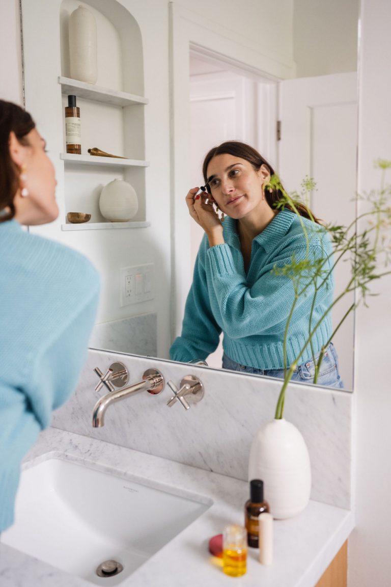 Brunette woman applying mascara in bathroom mirror.