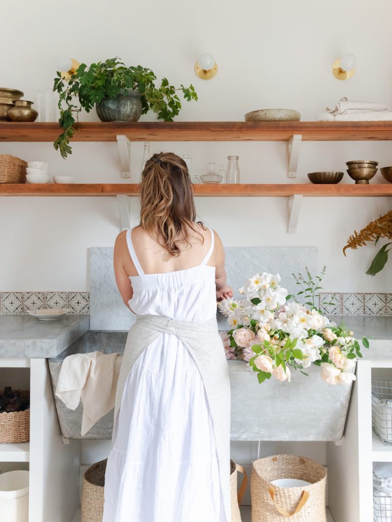Woman wearing white dress arranging flowers at sink.