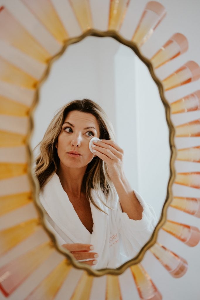 Blonde woman applying makeup in mirror.