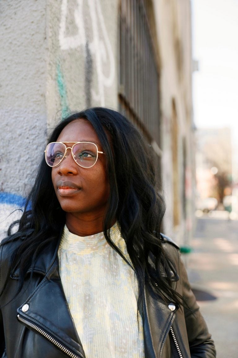 Black woman wearing glasses and leather jacket standing outside.