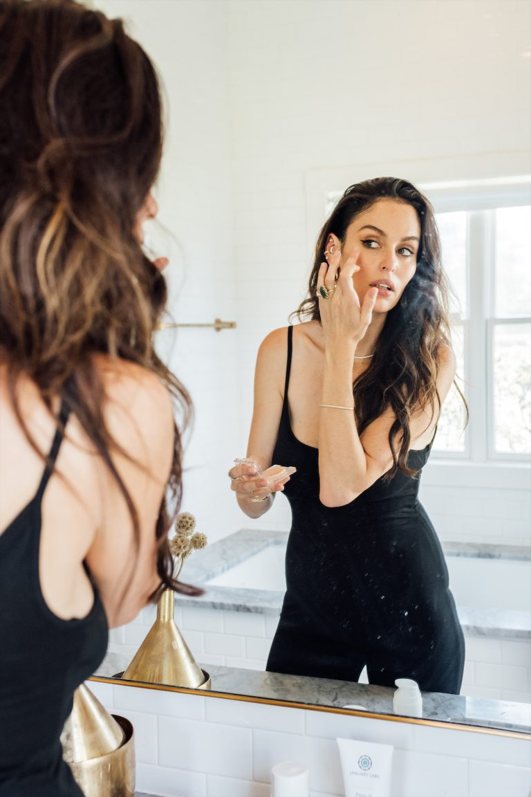 Woman applying skincare to face in mirror.