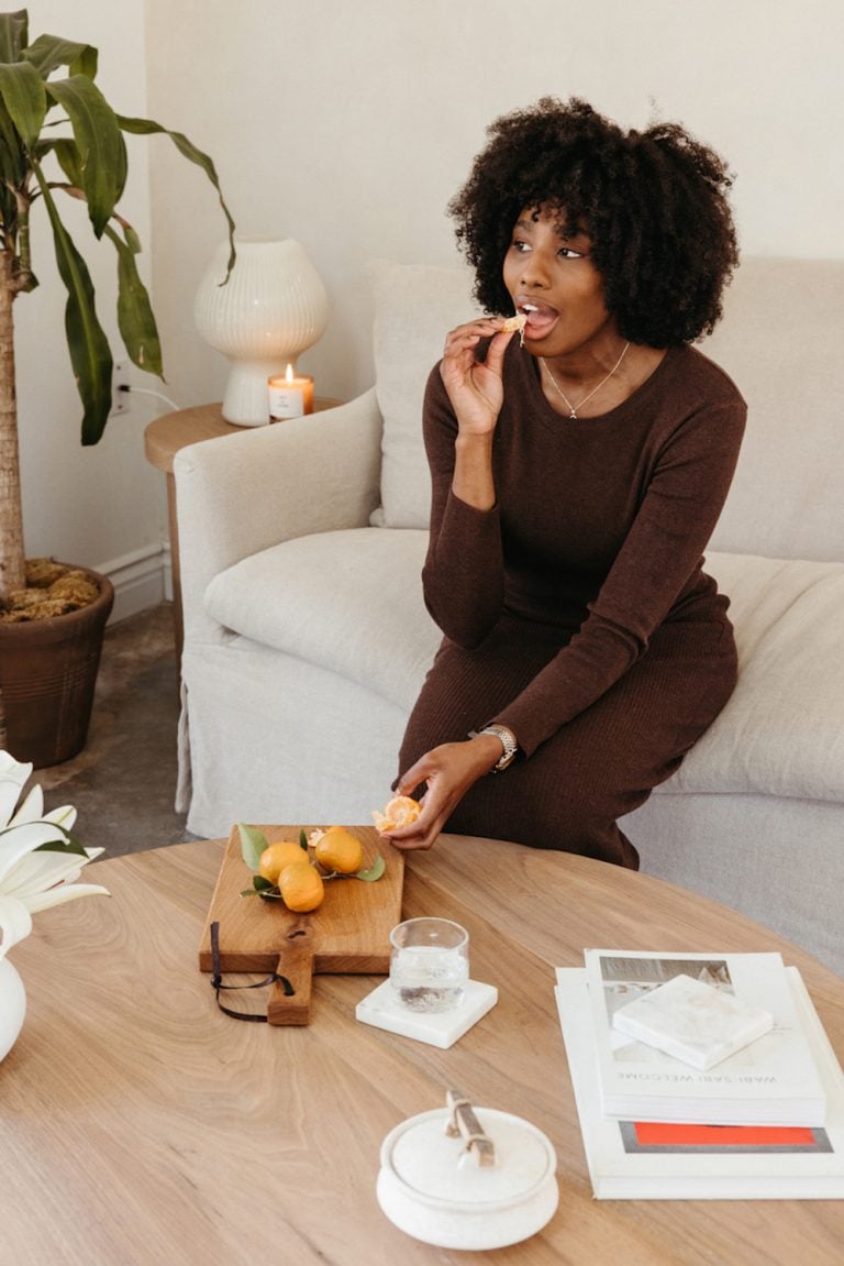 A black woman sitting on a sofa and eating an orange piece.