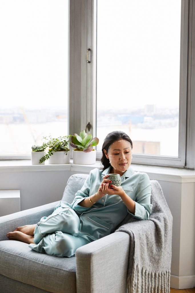 Woman drinking tea in armchair wearing pajamas and hydrogel mask.