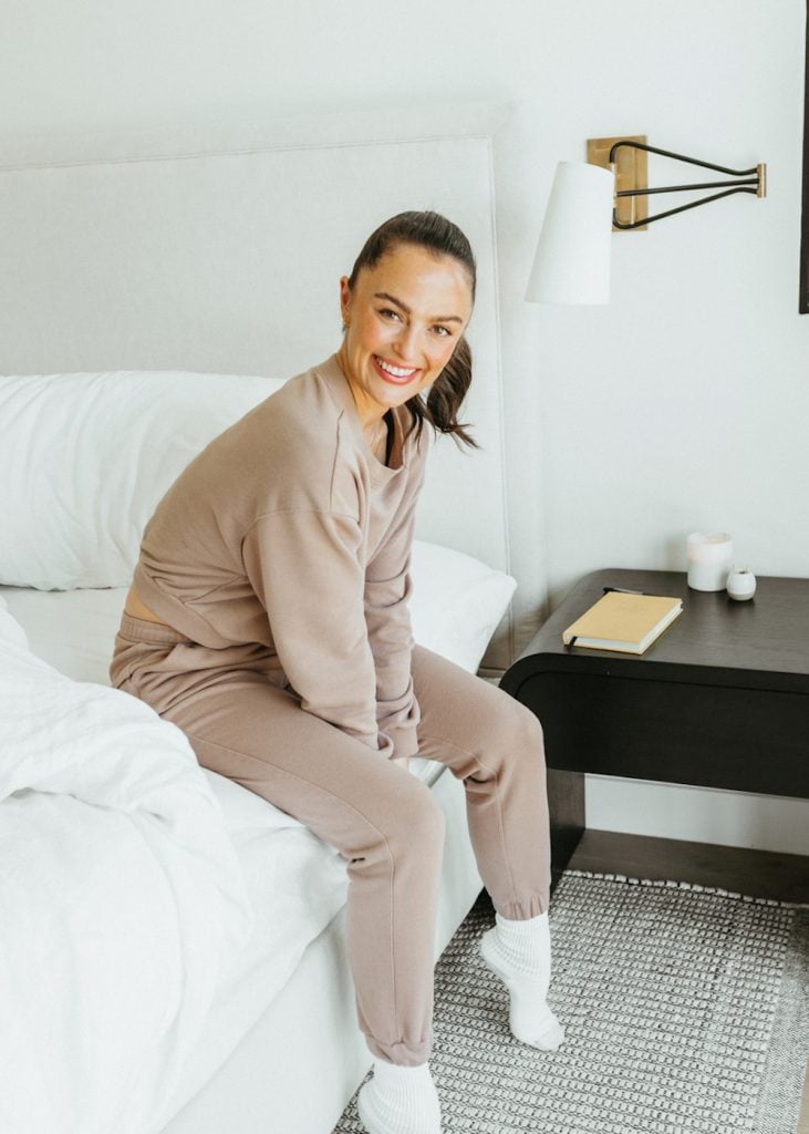 Megan Roup wearing cozy sweatsuit sitting on bed.