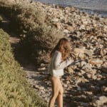 Woman walking on beach wearing swimsuit and drinking coffee.