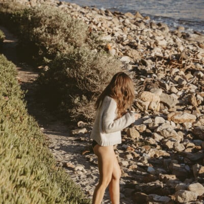 Woman walking on beach wearing swimsuit and drinking coffee.