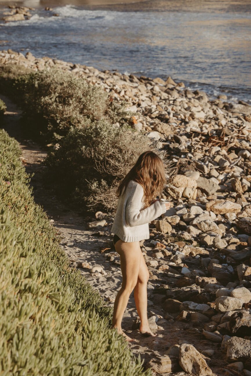 Woman walking on rocky beach holding coffee while taking a personal retreat.