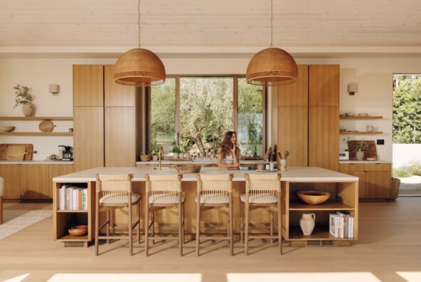 Camille Styles stands behind the oak kitchen island in her Malibu home, with woven pendant lights and wood counter stools.