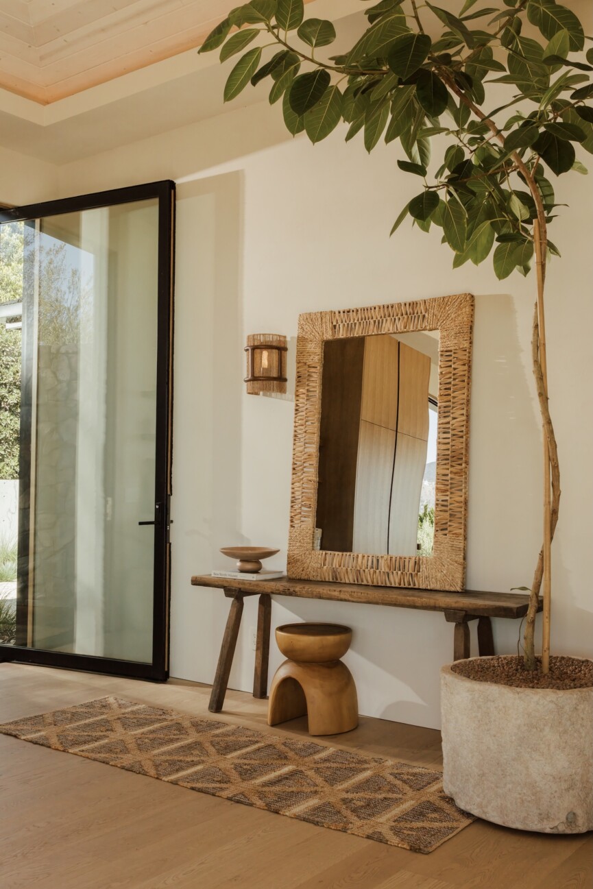 Sunlit entryway with a woven mirror, indoor tree, and doors open.