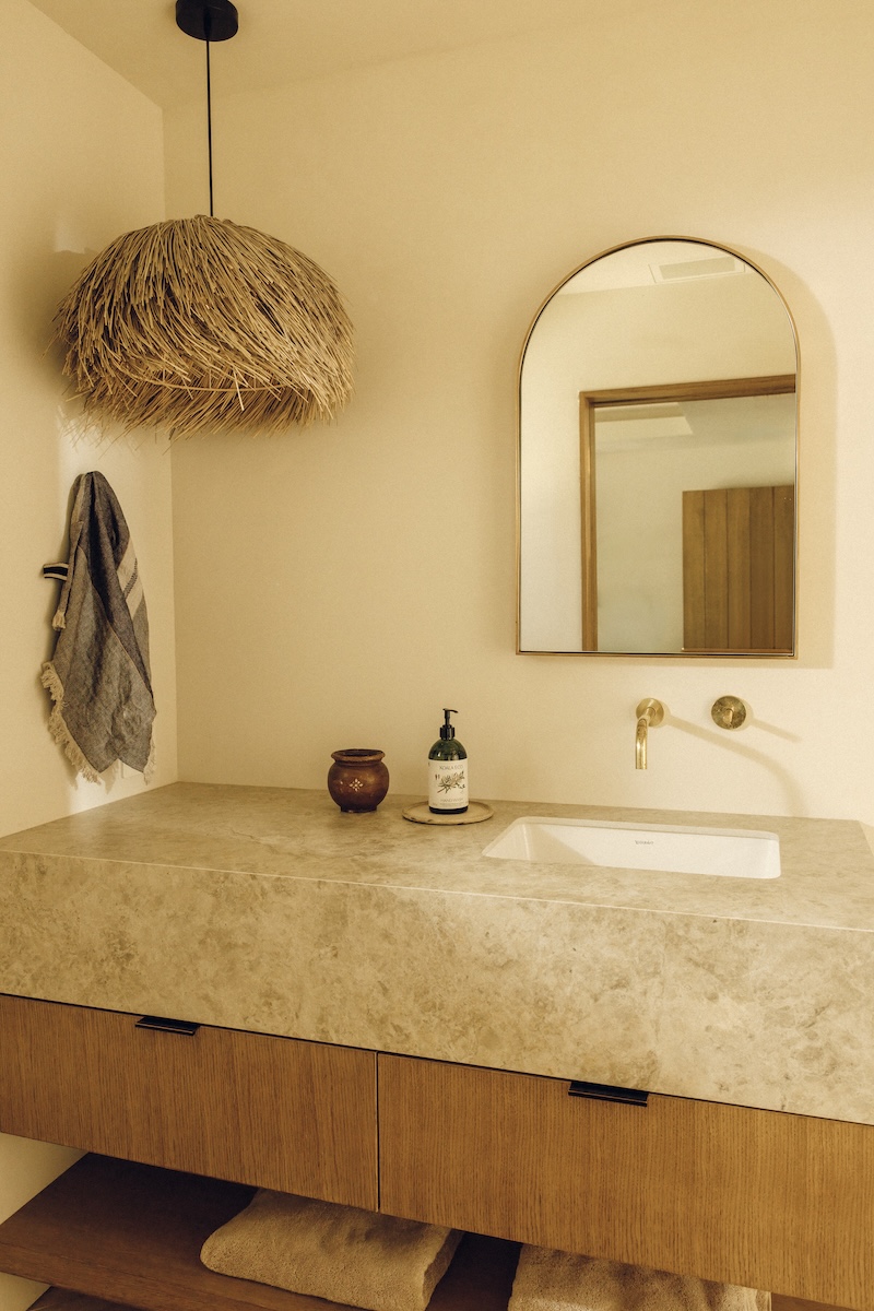 Boys’ bathroom vanity with warm wood drawers, a stone countertop, arched mirror, and woven pendant light.