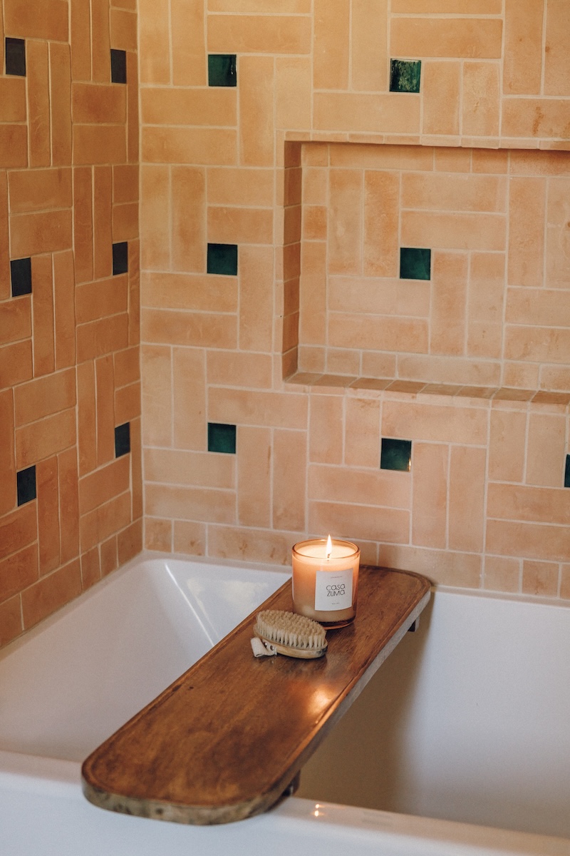 White bathtub with a live-edge wood bath tray against peach and green tiled walls.