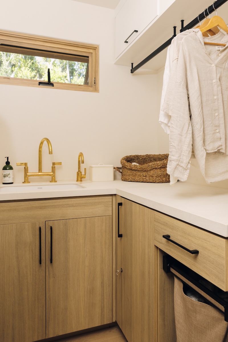 Laundry room utility sink with a brass faucet, wood cabinetry, woven basket, and wall-mounted drying rail.