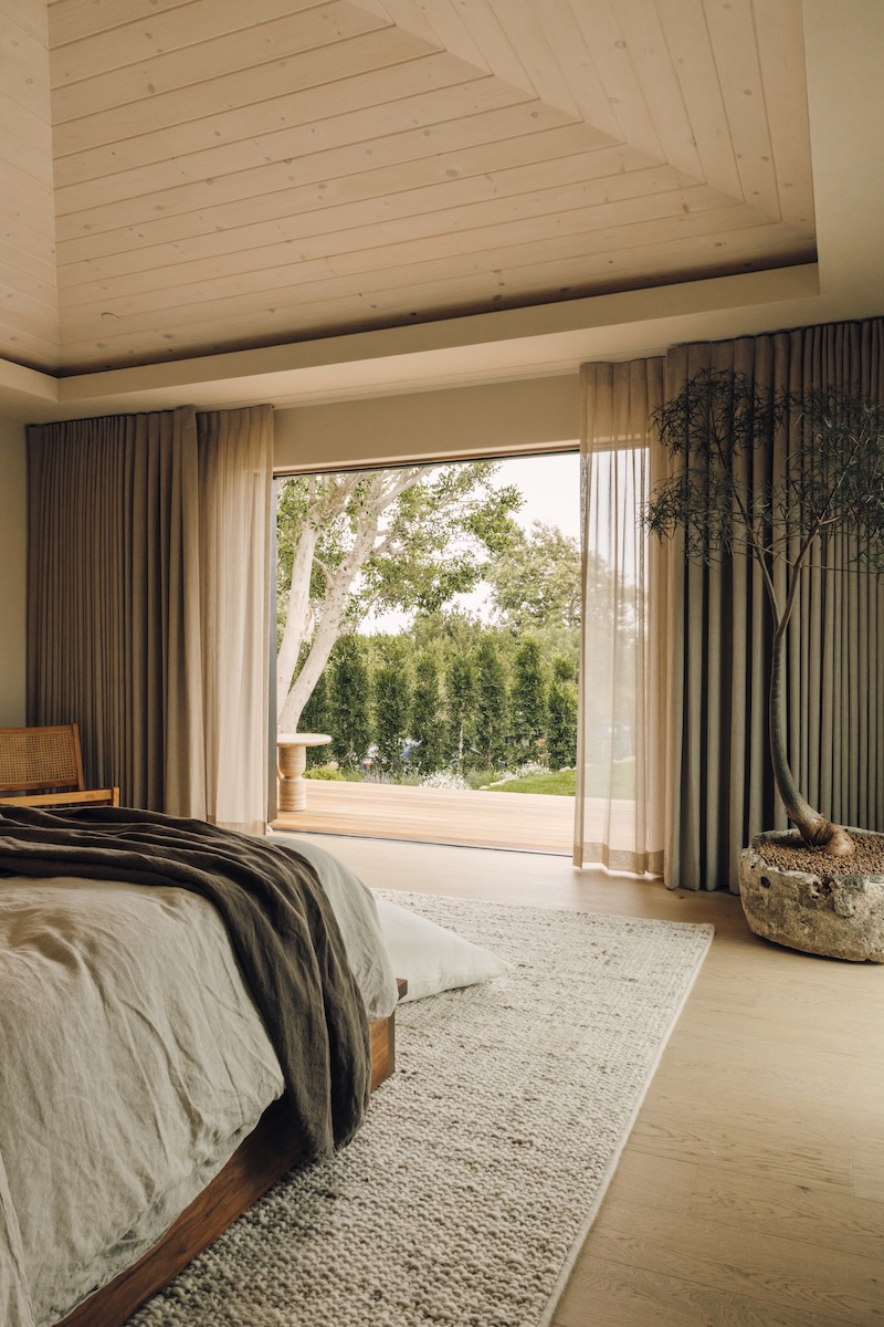 Malibu bedroom with a neutral bed, layered curtains, and an open doorway overlooking the garden.