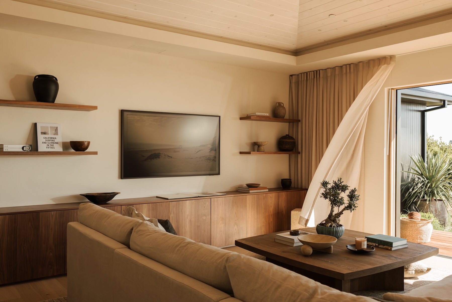 Camille Styles’ Malibu family room with a neutral sectional, wood media cabinet, floating shelves, and open garden doors.