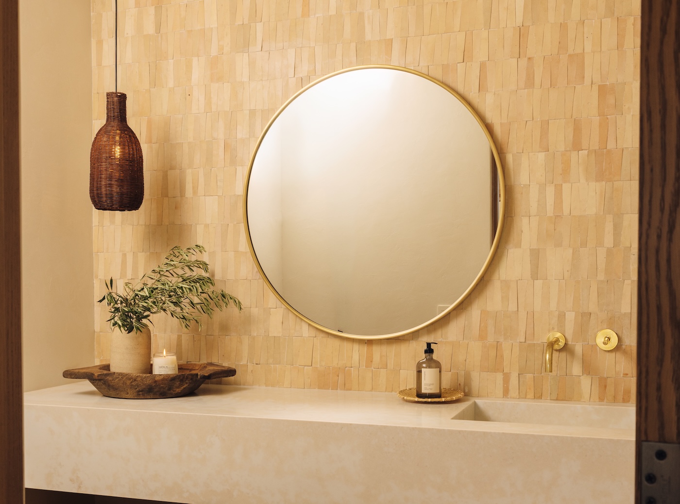 Camille Styles’ Malibu powder bathroom with warm tiled walls, a floating stone vanity, round mirror, and amber pendant light.