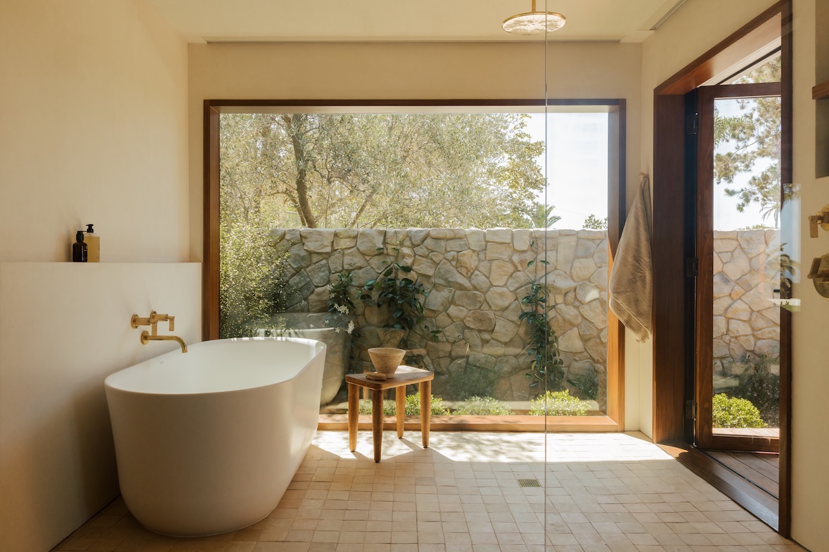 Camille Styles’ Malibu primary bathroom with a freestanding tub, brass fixtures, and floor-to-ceiling garden views.
