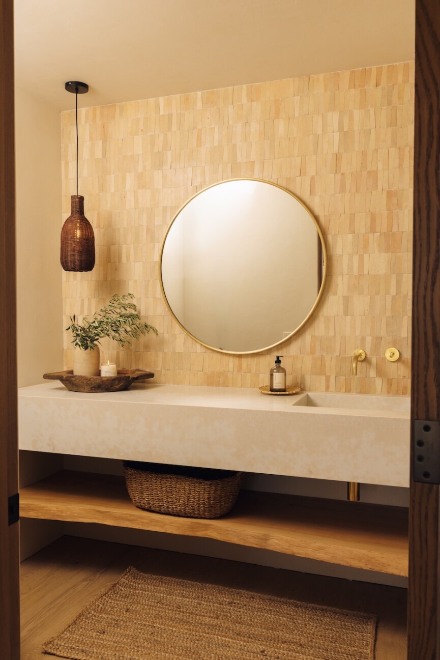 Powder bathroom vanity with warm tile, a round mirror, integrated sink, reclaimed wood shelf, and wall-mounted brass fixtures.